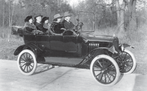 Imagem: Fotografia em preto e branco. Cinco pessoas estão sentadas dentro de um carro antigo. Ao fundo, árvores.   Fim da imagem.