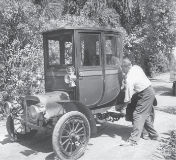 Imagem: Fotografia em preto e branco. Um homem está girando uma manivela na lateral de um carro antigo. Em volta há plantas e árvores.   Fim da imagem.