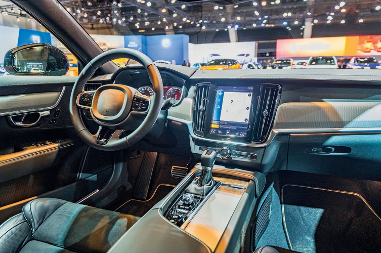 Imagem: Fotografia. Interior de um carro com volante e assentos de couro. Ao lado do volante há um painel grande com um mapa.   Fim da imagem.