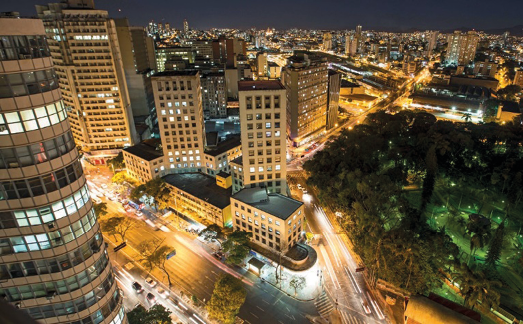 Imagem: Fotografia. Vista aérea de uma cidade a noite. No centro há carros com os faróis acesos em ruas e nas laterais, prédios com as luzes acesas e árvores.  Fim da imagem.