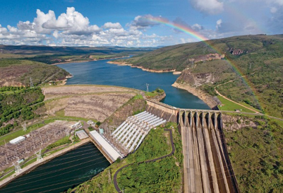 Imagem: Fotografia. Vista aérea de uma barragem ao lado de um lago. Ao fundo, morros e árvores.  Fim da imagem.