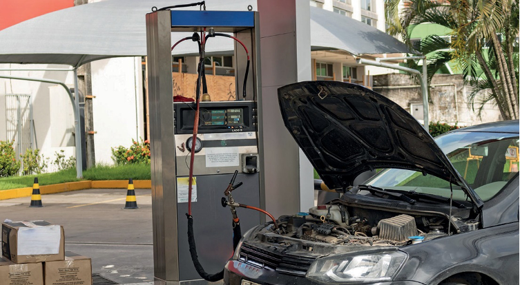 Imagem: Fotografia. Um carro parado ao lado de uma bomba de gasolina. O capô do carro está aberto e há um cabo conectado nele.  Fim da imagem.