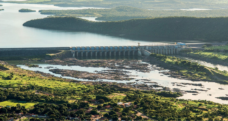 Imagem: Fotografia. Vista aérea de uma barragem ao lado de um lago grande. Em volta há morros e árvores. Fim da imagem.