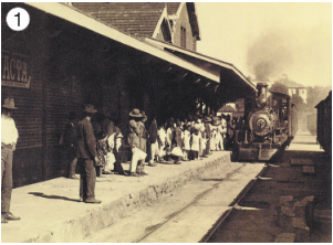 Imagem: Fotografia 1 em preto e branco. Pessoas paradas em uma plataforma. Ao lado, um trem se aproxima sobre trilhos.  Fim da imagem.