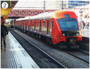 Imagem: Fotografia 2. À esquerda, pessoas em uma plataforma. No centro, um trem laranja sobre trilhos.   Fim da imagem.