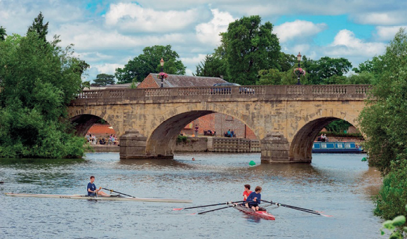 Imagem: Fotografia. Pessoas estão sentadas em barcos e segurando remos sobre um rio. Ao fundo, uma ponte e nas laterais, várias árvores. Fim da imagem.