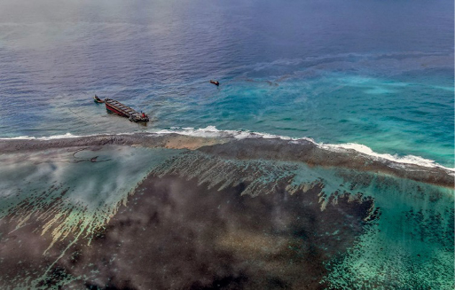 Imagem: Fotografia. Vista aérea de uma embarcação afundando no mar em tons de verde. Ao lado há uma mancha grande e preta na água.  Fim da imagem.