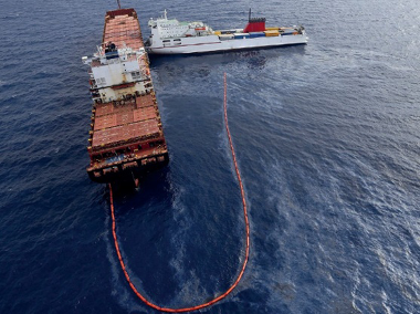 Imagem: Fotografia. Vista aérea de uma embarcação com uma corda amarrada na parte de trás. A outra ponta da corda está amarrada em um barco ao lado. E entre eles há uma mancha preta no mar.  Fim da imagem.