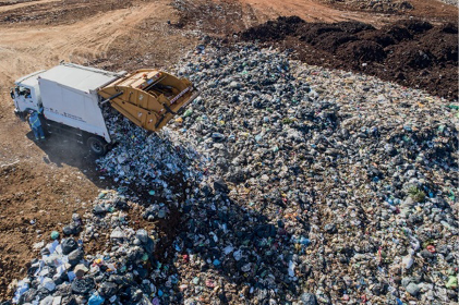 Imagem: Fotografia. Vista aérea de um caminhão despejando lixo sobre um aterro sanitário. Fim da imagem.