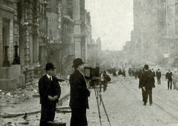 Imagem: Fotografia em preto e branco. Dois homens com chapéu e terno estão em pé, atrás de uma câmera fotográfica, que está em cima de um tripé. Ao fundo há homens andando em uma rua e nas laterais há prédios e fumaça.  Fim da imagem.