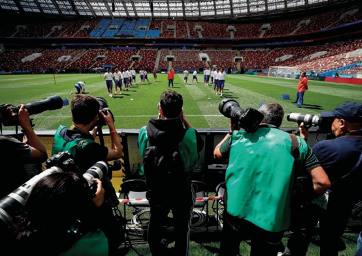 Imagem: Fotografia. Vários fotógrafos estão de costas e segurando câmeras na frente do rosto. Ao fundo, jogadores em um campo de futebol.  Fim da imagem.