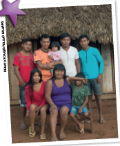 Imagem: Fotografia. Família indígena sentada em um banco em frente a uma oca. Na fileira à frente, menina de cabelo longo preto e camiseta vermelha, mulher de cabelo longo preto e camiseta roxa, menino de cabelo curto preto e camiseta verde. Na fileira inferior, menino de cabelo curto preto e camiseta vermelha, homem de cabelo curto castanho e camiseta laranja, segurando menina bebê de cabelo médio preto e camiseta rosa, menino de cabelo curto preto e camiseta branca, menino de cabelo curto preto e camiseta azul. Fim da imagem.