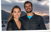 Imagem: Fotografia. Mulher de cabelo longo loiro, vestindo camiseta preta, está ao lado de um homem de cabelo curto e barba curta castanho, vestindo camiseta azul e casaco preto. Estão em frente ao mar e montanhas. Fim da imagem.