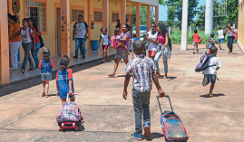 Imagem: Fotografia. Pátio com crianças andando segurando mochilas. Nas laterais, salas laranjas abertas com janelas grandes. Fim da imagem.