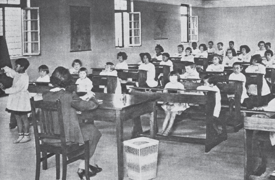 Imagem: Fotografia em preto e branco. Sala de aula com carteiras duplas com crianças sentadas e enfileiradas. À frente, mesa maior central com uma mulher. À esquerda, menina de cabelo curto, vestindo vestido, está em um quadro.  Fim da imagem.