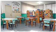 Imagem: Fotografia. Vista de sala com mesas quadradas com quatro cadeiras cada e prateleiras com livros. Fim da imagem.