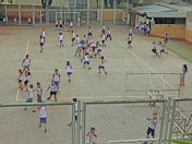 Imagem: Fotografia. Vista de quadra com traves de futebol e basquete e demarcações sobre o chão em faixas. Fim da imagem.