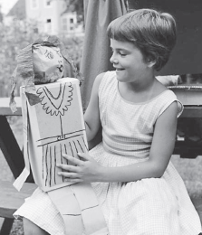 Imagem: Fotografia em preto e branco. Menina de cabelo curto, vestindo vestido curto. Está segurando uma boneca desenhada sobre papel encapando uma caixa. Fim da imagem.