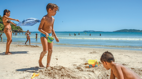 Imagem: Fotografia. Destaque de crianças sobre a areia de uma praia com brinquedos. Atrás há uma criança brincando com uma pipa e pessoas no mar. Fim da imagem.