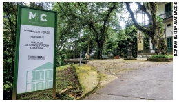 Imagem: Fotografia. Vista calçada de cimento e paralelepípedo com placa indicando “parque da cidade, preserve, unidade de conservação ambiental, MHC”. Atrás há bancos e árvores em uma praça, ao lado um casarão de dois andares branco e verde. Fim da imagem.