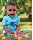 Imagem: Fotografia. Menino bebê de cabelo curto castanho, vestindo camiseta azul e bermuda branca. Está sentado em um campo de grama com brinquedos em blocos coloridos espalhados à frente. Fim da imagem.