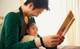 Imagem: Fotografia. Homem de cabelo curto castanho, vestindo camiseta verde. Está segurando um livro aberto, com um bebê no colo. Fim da imagem.