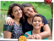 Imagem: Fotografia. Mulher de cabelo longo castanho e camiseta preta, abraçando menino de cabelo curto preto e camiseta azul, e menina de cabelo longo castanho e camiseta preta. Estão em frente a um lago e vegetação. Fim da imagem.