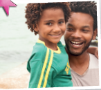 Imagem: Fotografia. Homem de cabelo curto castanho cacheado, vestindo camiseta branca, está segurando uma menina de cabelo curto castanho cacheado, vestindo camiseta verde. Fim da imagem.