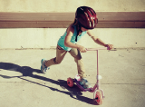 Imagem: Fotografia. Menina de cabelo longo e capacete vermelho, vestindo camiseta verde e calça marrom, está andando em um patinete rosa. Fim da imagem.