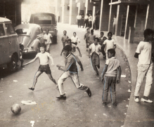 Imagem: Fotografia em preto e branco. Crianças em uma rua jogando uma bola. Ao redor há carros antigos estacionados. Fim da imagem.