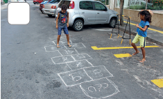 Imagem: Fotografia. Dois meninos pulando uma amarelinha desenhada sobre rua. Fim da imagem.