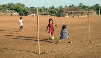 Imagem: Fotografia. Vista de campo de areia com duas traves. Há crianças jogando bola sobre o campo. Ao fundo há vegetação baixa e ocas de palha.  Fim da imagem.