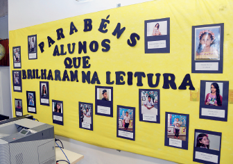 Imagem: Fotografia. Vista de quadro amarelo com fotografias de crianças expostas em retângulos azuis. No centro, o título: ”parabéns aos alunos que brilharam na leitura”. Fim da imagem.