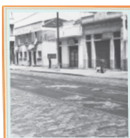 Imagem: Fotografia em preto e branco. Rua com lojas pequenas de porta de aço e placas pequenas.  Fim da imagem.