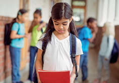 Imagem: Fotografia. Destaque de menina de cabelo longo preto, vestindo camiseta branca, segurando um livro vermelho. Atrás em imagem desfocada crianças conversam apontando para menina. Fim da imagem.