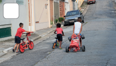 Imagem: Fotografia. Crianças segurando bicicletas pequenas e velotrol em uma ladeira. Fim da imagem.