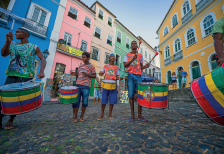 Imagem: Fotografia. Destaque de meninos com tambores pintados em faixa verde, amarela, azul e vermelha. Estão em uma ladeira de pedra com prédios baixos coloridos por toda a rua.  Fim da imagem.