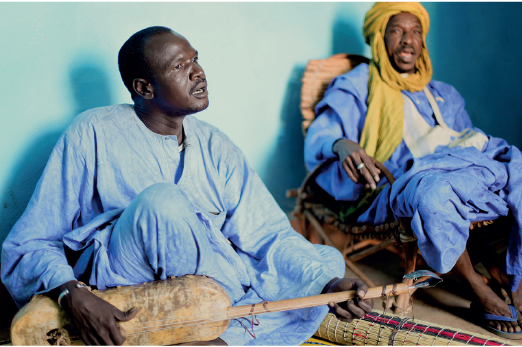 Imagem: Fotografia. Homem de cabelo curto preto, vestindo túnica azul. Está segurando um instrumento musical de madeira, sentado no chão. Ao lado, homem de turbante amarelo e túnica azul, sentado em uma cadeira de bambu. Fim da imagem.