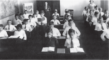 Imagem: Fotografia em preto e branco. Meninos sentados em carteiras duplas com livros abertos. Há três fileiras de carteiras. Fim da imagem.