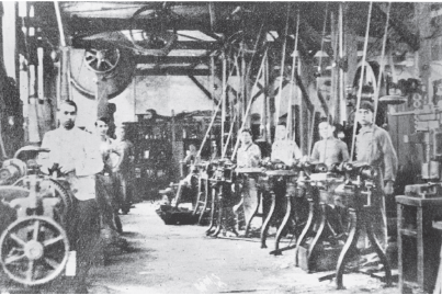 Imagem: Fotografia em preto e branco. Linha de produção com rodas, mesas ligadas por estruturas de ferro e cano. Há homens e meninos trabalhando ao redor das mesas. Fim da imagem.