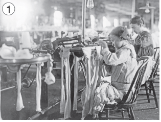 Imagem: Fotografia em preto e branco. Crianças sentadas em cadeiras em frente a máquina manual com tecidos longos sobre ela. Fim da imagem.