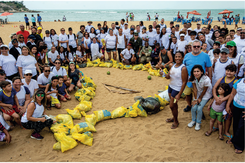 Imagem: Fotografia. Praia com pessoas reunidas ao redor de uma representação do território brasileiro formado por sacos de lixo. Fim da imagem.