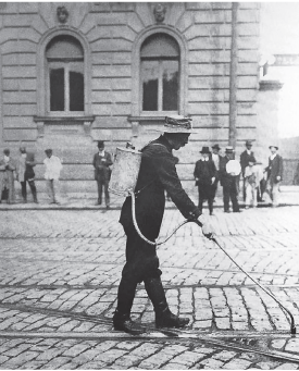 Imagem: Fotografia em preto e branco. Homem de chapéu longo, vestindo casaco e calça. Está segurando um pequeno reservatório nas costas ligado a um cano longo que passa em trilho de um bonde. Fim da imagem.