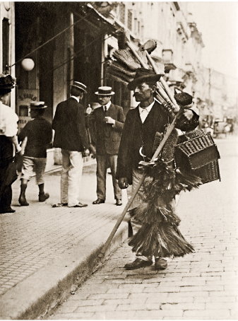 Imagem: Fotografia em preto e branco. Homem de chapéu, cabelo curto e bigode, vestindo camisa, casaco e calça. Está levando no ombro vassouras com cerdas longas, espanadores e cestas.  Fim da imagem.