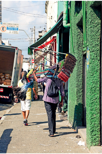 Imagem: Fotografia. Homem de cabelo curto, vestindo camiseta lilás e calça preta. Leva no ombro vassouras coloridas com cerdas pequenas. Fim da imagem.