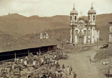 Imagem: Fotografia em preto e branco. Vista de igreja com torres laterais e faixada larga com cruz ao topo, sobre campo aberto de terra. À frente da igreja, um mercado aberto baixo com charretes e cavalos. Há pessoas na estrada de terra. Fim da imagem.