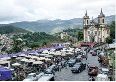 Imagem: Fotografia. Vista de igreja branca com acabamento bege, possui torres laterais e faixada larga com cruz ao topo. Ao redor, campo com árvores. À frente, barracas lado a lado, sobre a rua de paralelepípedo com carros. À esquerda, em segundo plano, morro com casas pequenas. Fim da imagem.