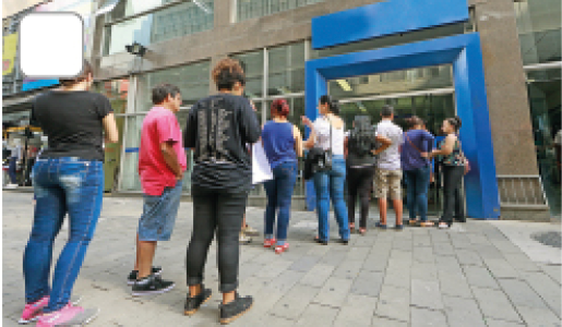 Imagem: Fotografia. Vista de pessoas em fila em frente a um banco com porta larga azul.  Fim da imagem.