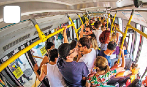 Imagem: Fotografia. Vista de ônibus lotado com pessoas em pé segurando nos suportes amarelos. Fim da imagem.