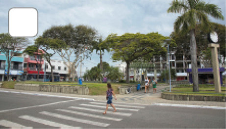 Imagem: Fotografia. Praça com vegetação e calçada larga. À frente, há uma mulher andando em uma faixa de pedestres na rua. Fim da imagem.